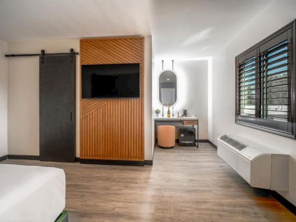 The image shows a modern hotel room with a bed, wooden wall paneling, TV, mirror, vanity desk, chair, and window with blinds.