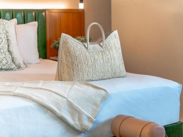 A neatly made bed with a patterned cushion, a wicker bag, a cozy throw, and a rolled-up towel on top of a white sheet.