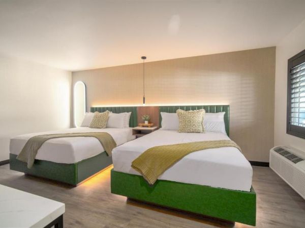 A hotel room with two neatly made beds, green accents, modern decor, and a window with shutters on the right side.