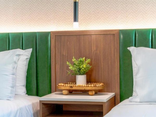 A bedside table with a small plant and a decorative tray is flanked by two beds with green headboards and white pillows.