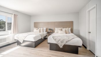 The image shows a minimalistic bedroom with two beds, light wood flooring, a window, and neutral curtains, decorated in a modern style.
