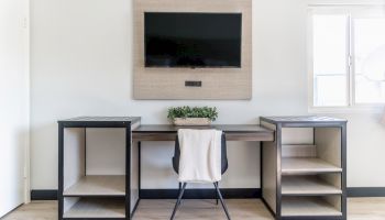 A modern workspace with a TV, desk, chair, shelves, and plant under a window. The setup is minimal and neatly arranged.