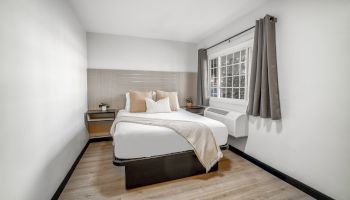 A modern bedroom with a neatly made bed, decorative pillows, and nightstands, illuminated by natural light from a window with gray curtains.
