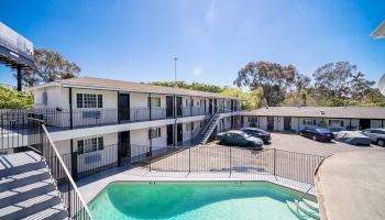 The image shows a two-story motel with a parking lot and a fenced swimming pool under a clear blue sky.
