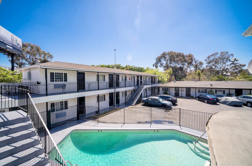 The image shows a two-story motel with a parking lot and a fenced swimming pool under a clear blue sky.