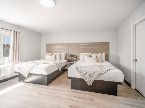 The image shows a bright, modern bedroom with two neatly made beds, wooden flooring, a window with curtains, and a small table between the beds.