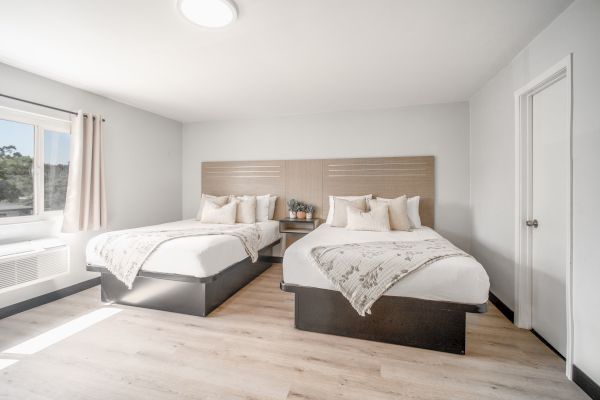 The image shows a bright bedroom with two double beds, decorative pillows, a small table, and a window with curtains.