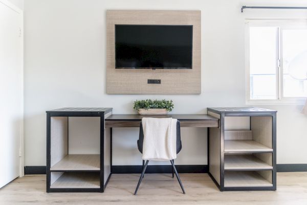 The image shows a minimalist desk setup with a chair, a wall-mounted TV, shelves, and a small plant. Natural light fills the room.