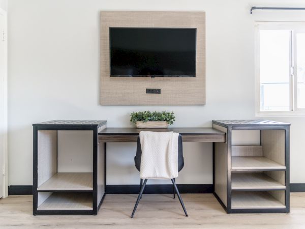 The image shows a minimalist desk setup with a chair, a wall-mounted TV, shelves, and a small plant. Natural light fills the room.