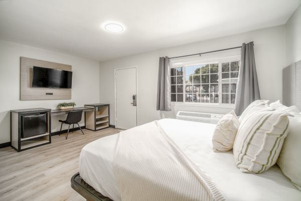 The image shows a bright bedroom with a bed, TV, desk, chair, window, and curtains, creating a modern and minimalistic atmosphere.