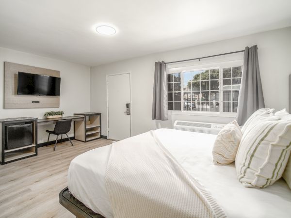The image shows a bright bedroom with a bed, TV, desk, chair, window, and curtains, creating a modern and minimalistic atmosphere.