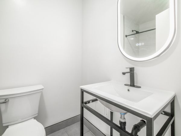 The image shows a modern bathroom with a toilet, a sink on an open metal-framed vanity, and an illuminated mirror above the sink.