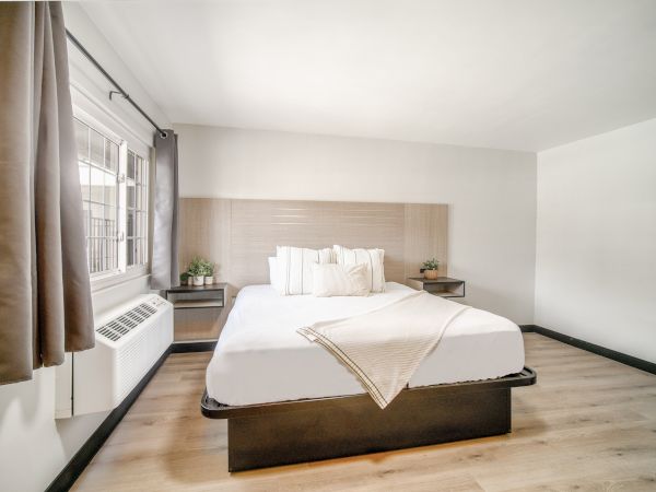 A minimalistic bedroom with a neatly made bed, pillows, and a window to the left, featuring neutral colors and simple decor.