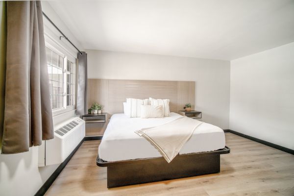 A minimalistic bedroom with a neatly made bed, wooden floor, window with curtains, and two small side tables with plants on them.