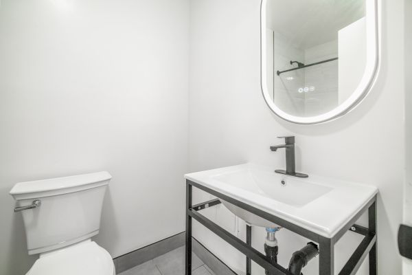 A minimalist bathroom features a toilet, a modern sink with a black faucet, and an illuminated oval mirror.