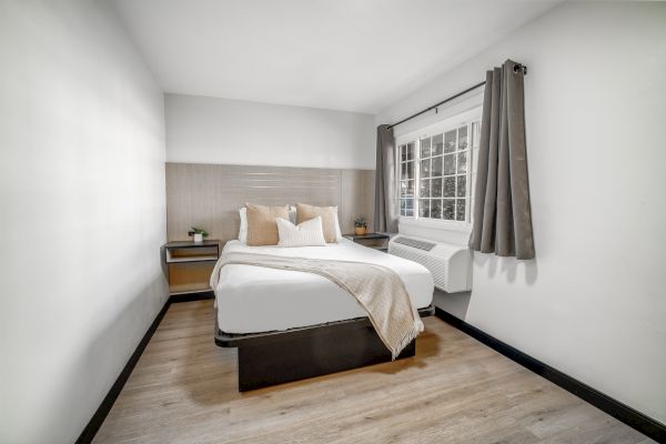 A minimalist bedroom with a neatly made bed, pillows, a blanket, nightstands, and curtains, featuring wooden flooring and a window.
