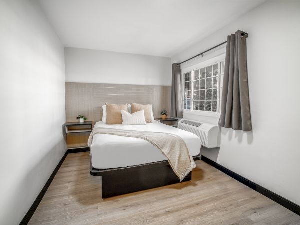 A minimalist bedroom with a neatly made bed, pillows, a blanket, nightstands, and curtains, featuring wooden flooring and a window.