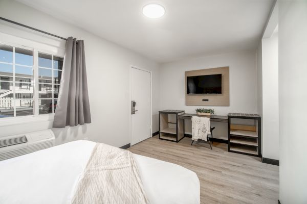 A minimalistic room with a bed, desk, and TV on the wall, featuring light decor and a window with grey curtains.