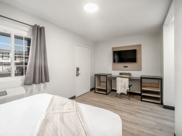 A minimalistic room with a bed, desk, and TV on the wall, featuring light decor and a window with grey curtains.