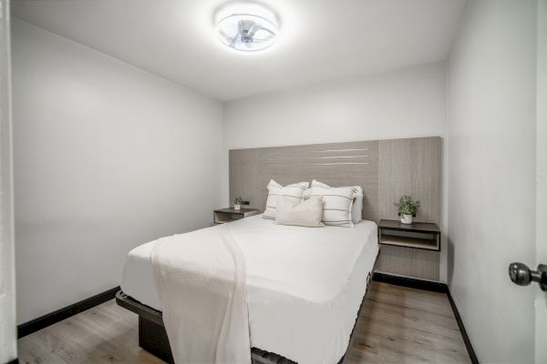 A small, minimalist bedroom with a bed, white linens, two side tables, and a ceiling light, set in a clean, modern design.
