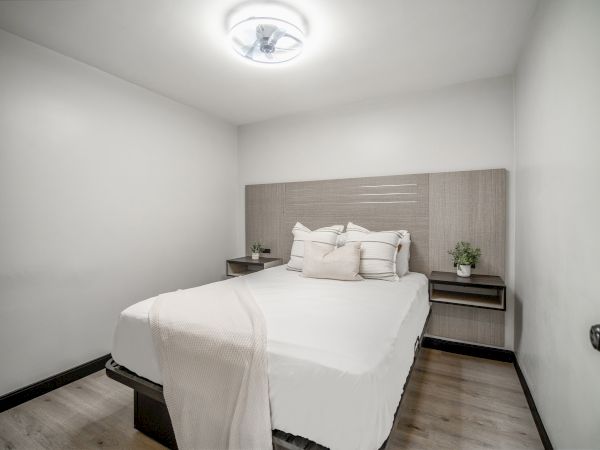 A small, minimalist bedroom with a bed, white linens, two side tables, and a ceiling light, set in a clean, modern design.