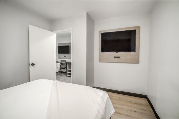A minimalist bedroom with white walls, a bed, a wall-mounted TV, and an open door leading to another room with a desk and chair.