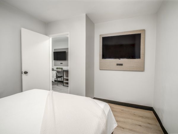 A minimalist bedroom with white walls, a bed, a wall-mounted TV, and an open door leading to another room with a desk and chair.