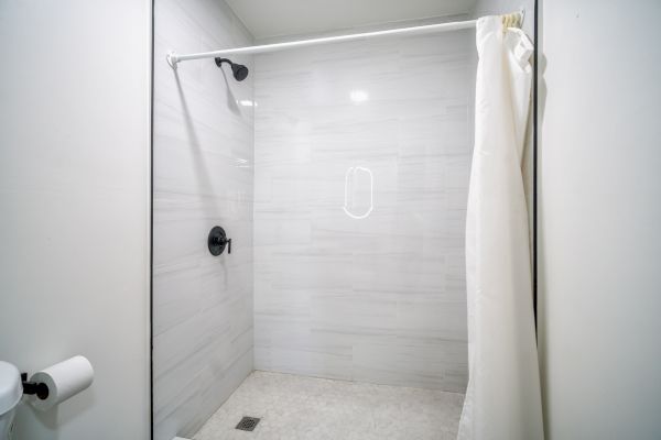 The image shows a modern shower with light gray tiles, a black showerhead, a white curtain, and a white wall-mounted toilet paper holder.