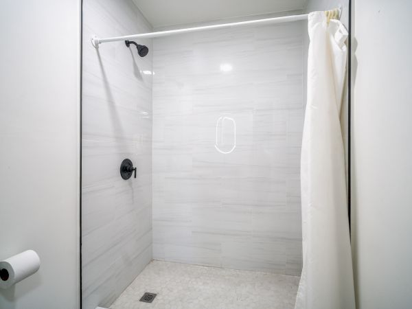 The image shows a modern shower with light gray tiles, a black showerhead, a white curtain, and a white wall-mounted toilet paper holder.