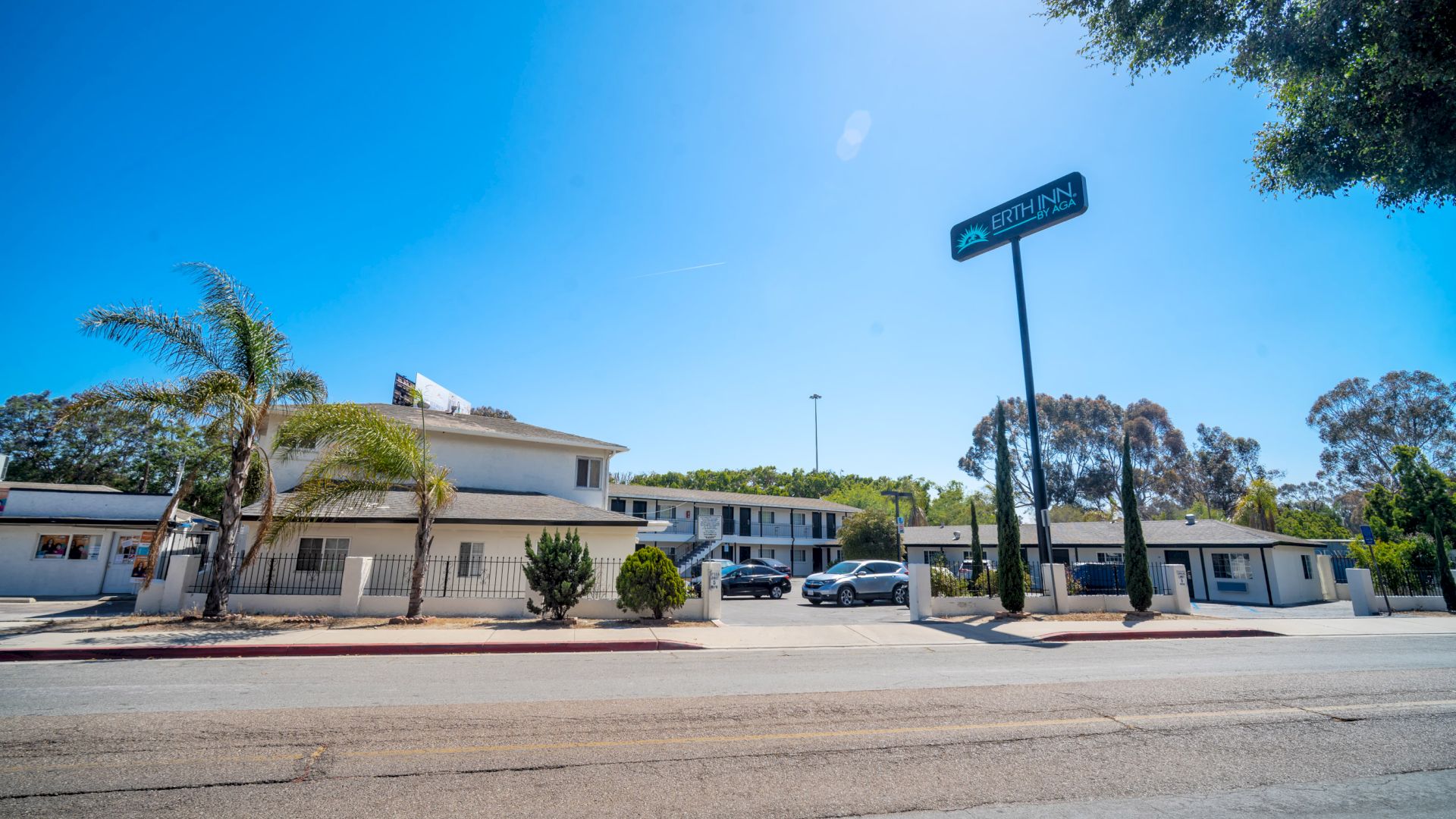 A roadside motel with a sign, several parked cars, palm trees, and a clear blue sky.