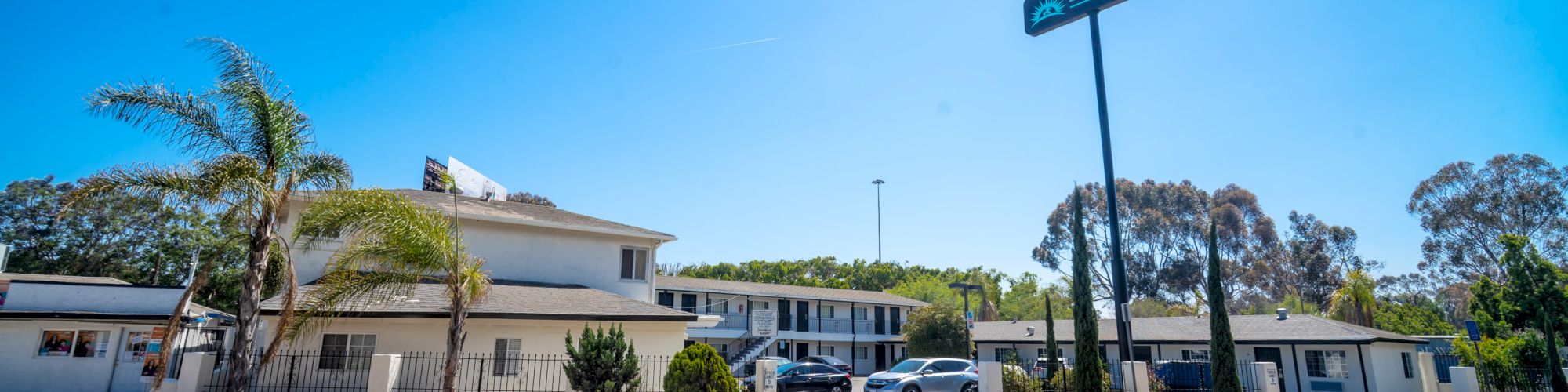 A roadside motel with a sign, several parked cars, palm trees, and a clear blue sky.