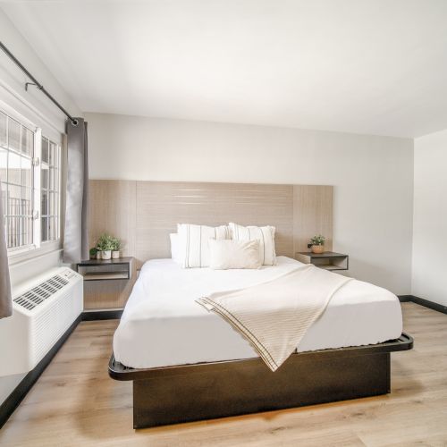 A minimalist bedroom with a neatly made bed, light wood flooring, side tables, and a window with a white curtain.