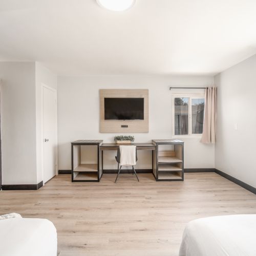 The image shows a minimalist room with a TV, desk, shelves, large windows, and two beds with white linens on either side.