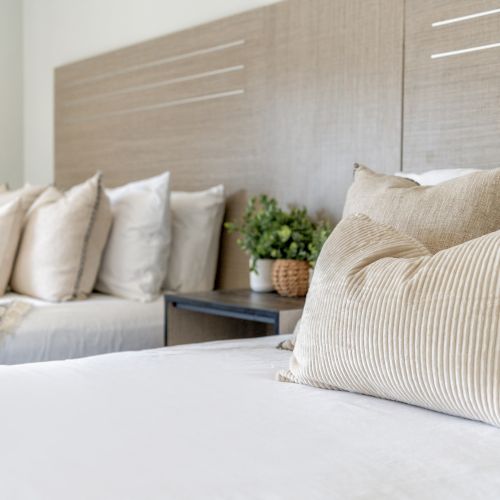 The image shows two neatly made beds with light-colored pillows and a wooden headboard, separated by a nightstand holding potted plants.