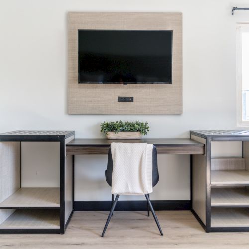 A minimalist workspace featuring a desk, a chair with a draped blanket, open shelves, a wall-mounted TV, and a window to the right.