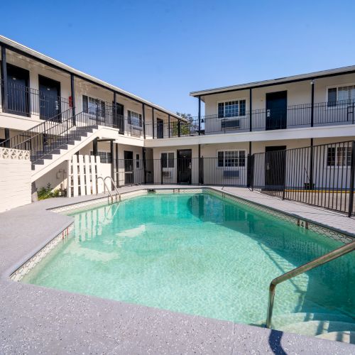 A courtyard with a pool is surrounded by a two-story building, featuring railings and stairs under a clear blue sky.