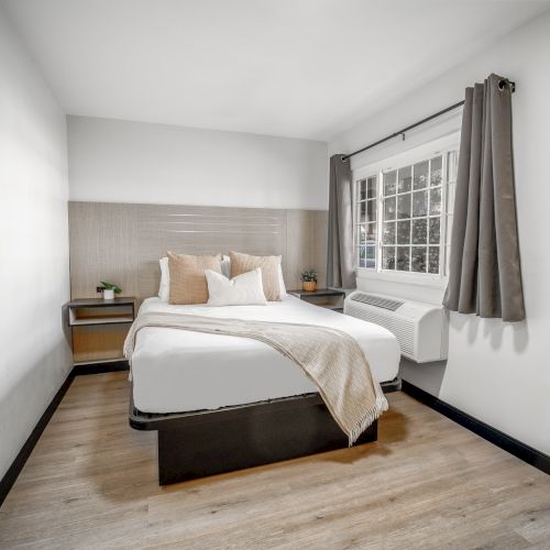 A modern bedroom with a neatly made bed, two pillows, a light blanket, bedside tables, a window with curtains, and wooden flooring.