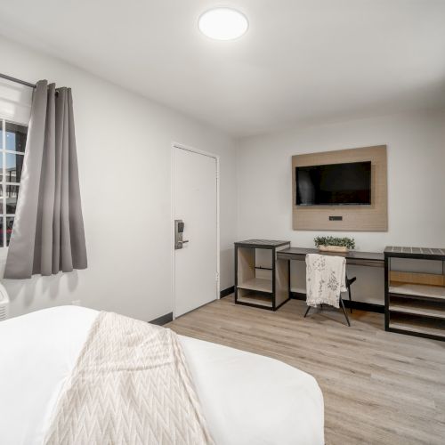 A modern room with a bed, window, desk, chair, wall-mounted TV, and light wood flooring, featuring neutral tones and minimal decor.