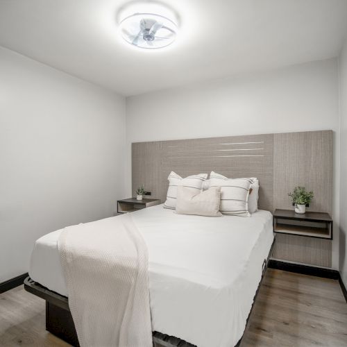 A modern bedroom with a bed, pillows, blanket, two side tables, and a plant, featuring minimalist decor and neutral tones.