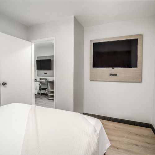 The image shows a minimalistic bedroom with a bed, a flat-screen TV on the wall, and an open door leading to another room with a desk.
