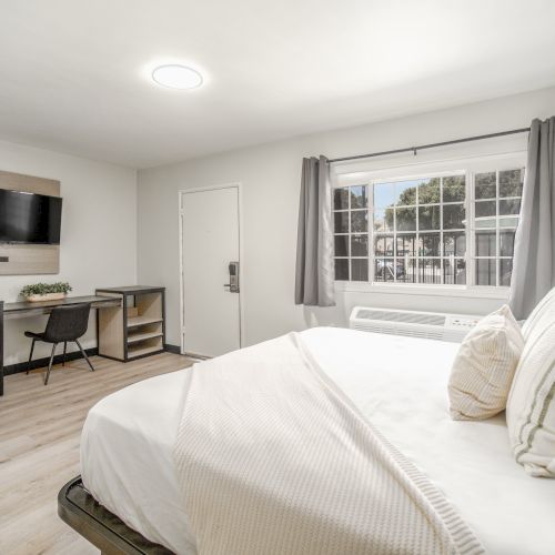 A modern, bright bedroom with a neatly made bed, desk, chair, TV, and window with curtains, offering a clean, minimalist look.