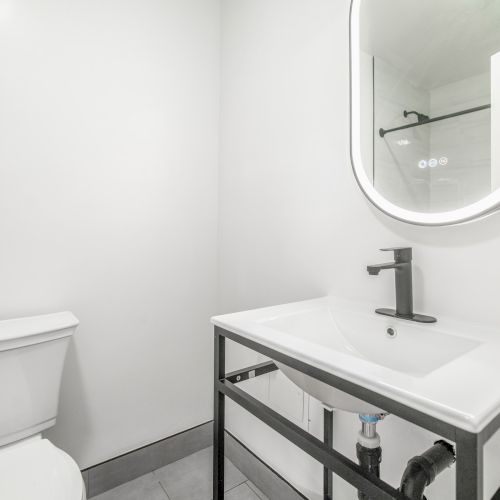 A modern bathroom with a toilet, rectangular sink on a metal frame, and a backlit oval mirror on a white wall.