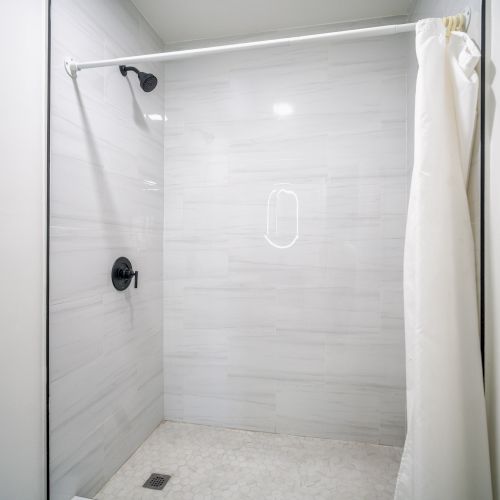 The image shows a white tiled shower area with a curtain, a black showerhead, and a toilet paper holder on the side.