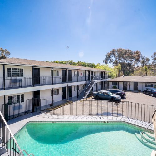The image shows a motel with outdoor stairs, a parking area, and cars. A swimming pool is in the foreground, surrounded by a fence.