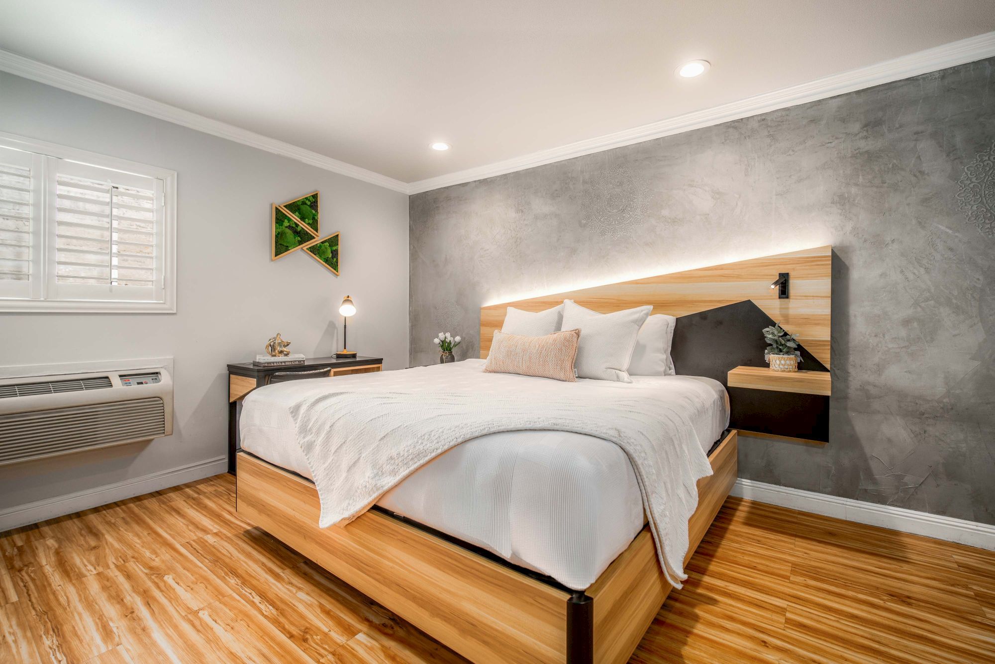 A modern bedroom with a wooden bed and headboard, light bedding, a small nightstand, window with blinds, and a textured gray accent wall.