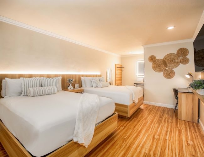 A bright hotel room with two queen beds, light wood furniture, a desk, wall art, and a mounted TV; warm wood flooring and soft lighting.