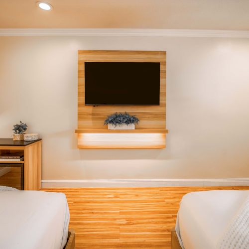 A hotel-style bedroom with two white beds, a wooden floor, a wall-mounted TV framed by wood, a shelf with a plant, and a small desk with a lamp on the left.