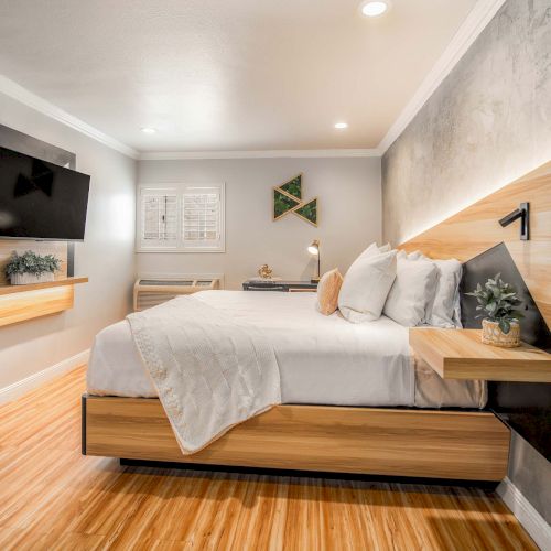 A modern hotel-style bedroom with a wooden platform bed, gray walls, a mounted TV, floating nightstands, and warm wood flooring, clean and bright.