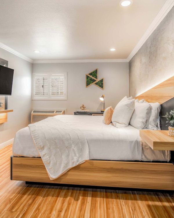 A modern hotel-style bedroom with a wooden platform bed, gray walls, a mounted TV, floating nightstands, and warm wood flooring, clean and bright.