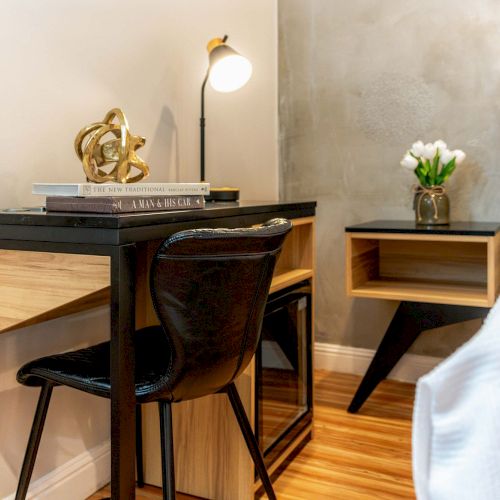 A tidy desk area beside a bed: a small black desk with a chair, books, a lamp, a decorative sculpture, and a nightstand with a plant on it.